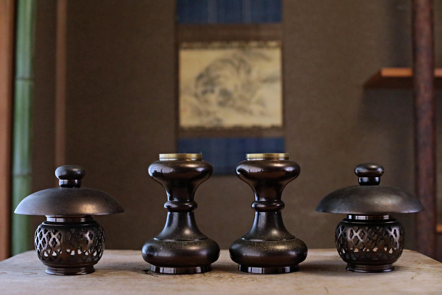 four dark brown, ornate candle holders arranged on a wooden surface, with a blurred background that includes a painting.