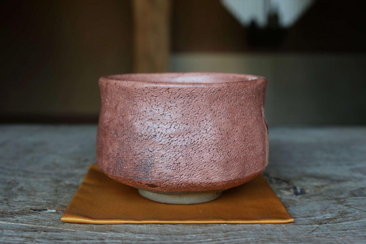 A close-up image of a pink ceramic bowl or teapot placed on a yellow cloth or mat on a wooden surface.