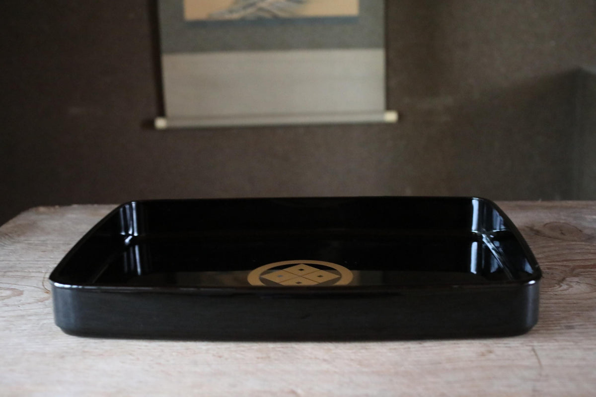 A black rectangular tray with a gold emblem on it, placed on a wooden surface.