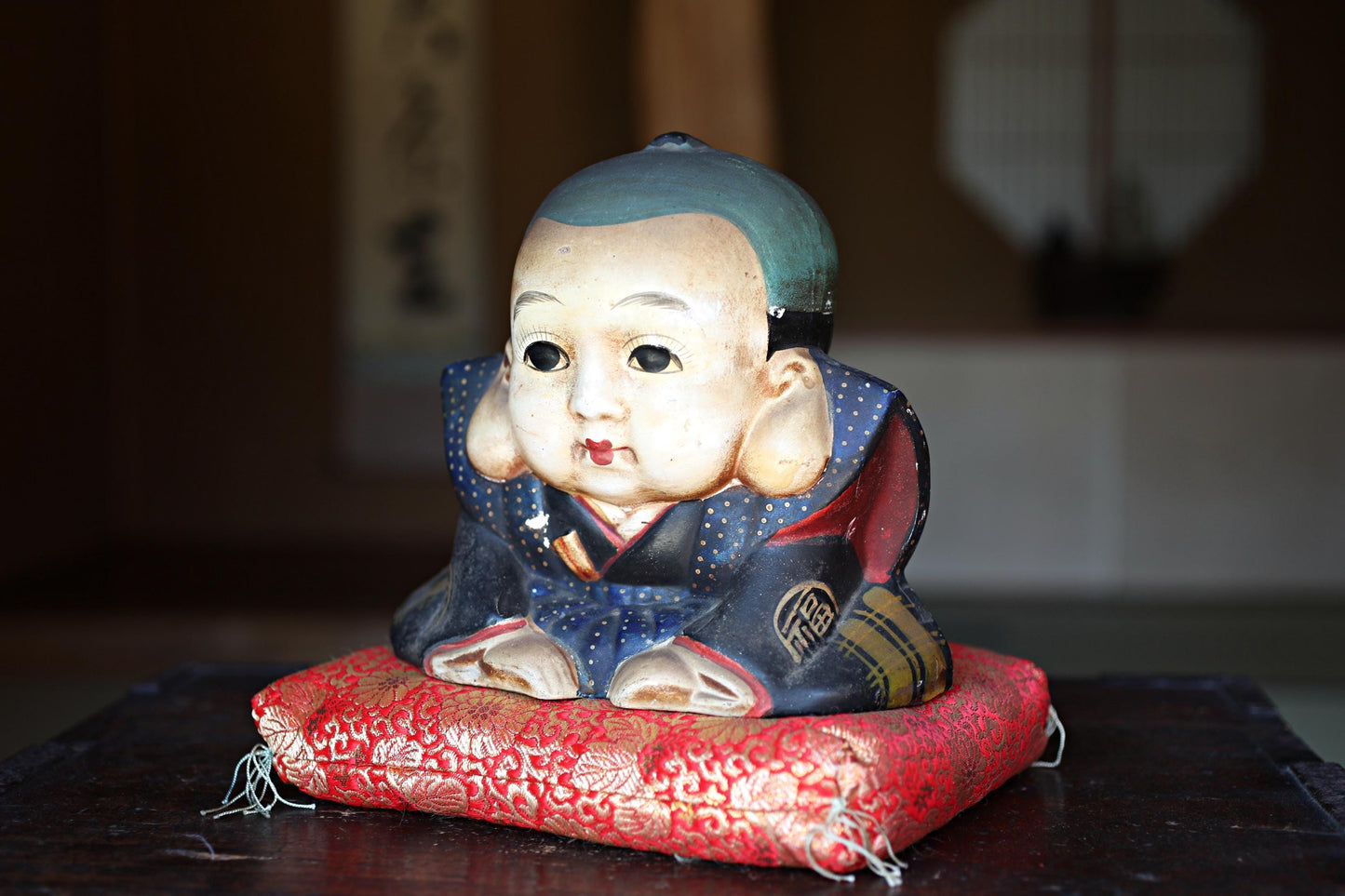 A ceramic figurine of a young boy sitting on a red cushion, with a traditional Japanese-style background.