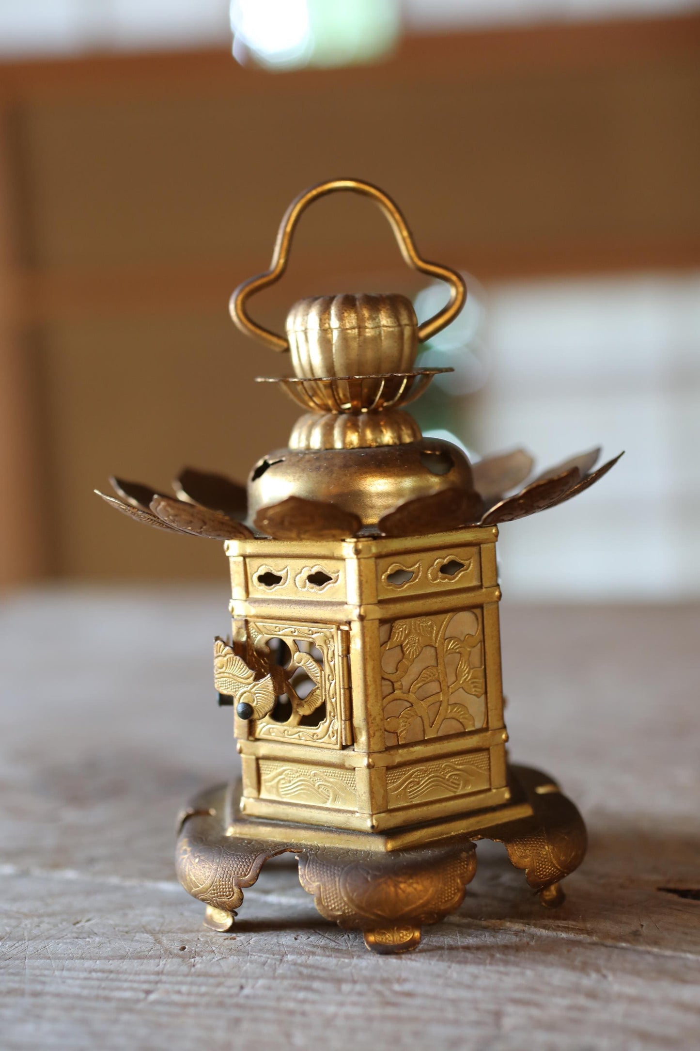 A small, ornate gold-colored lantern with a curved handle and intricate designs on the front sits on a wooden surface.