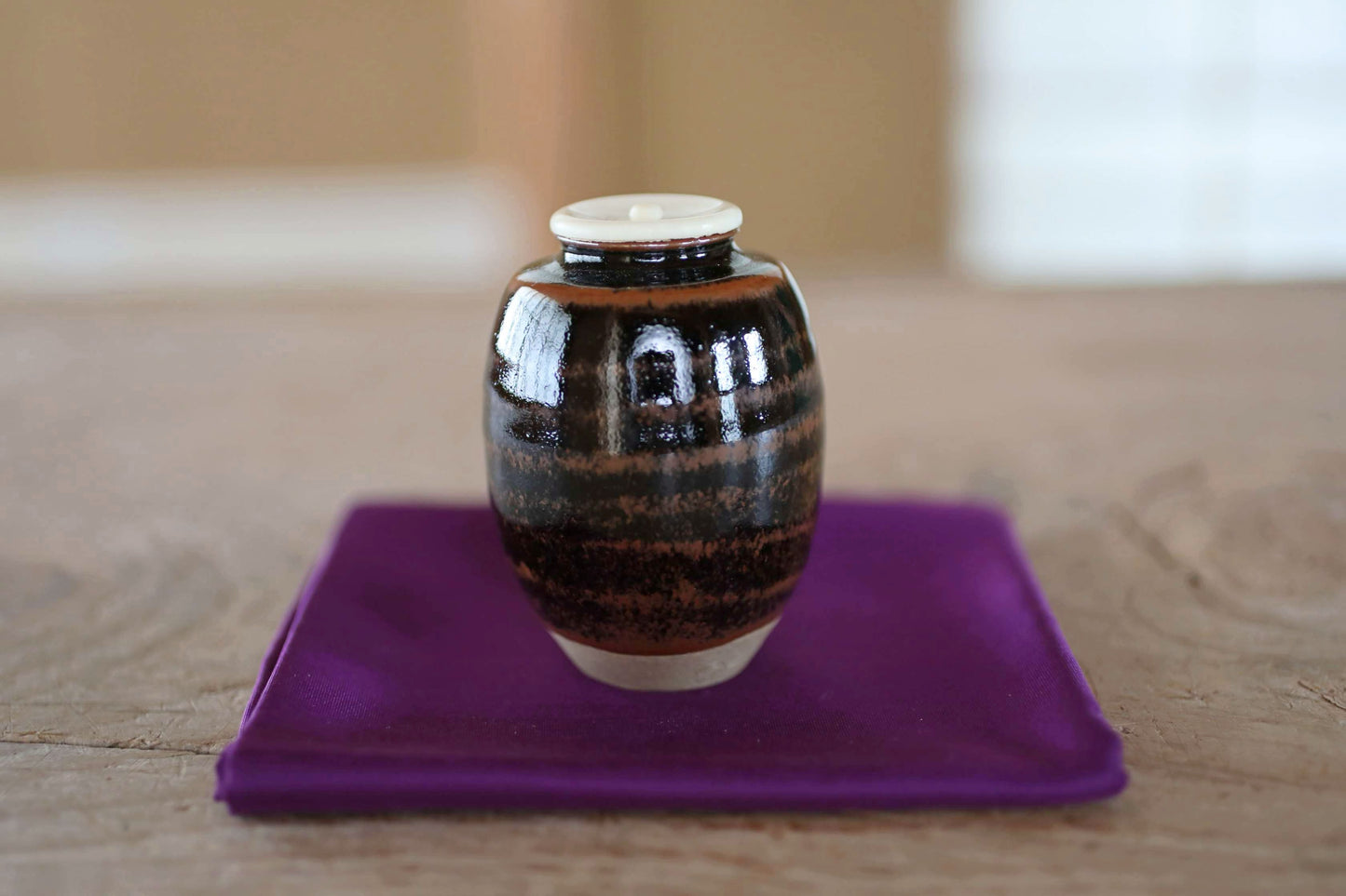 A small, round ceramic jar with a white lid sits on a purple cloth on a wooden surface.