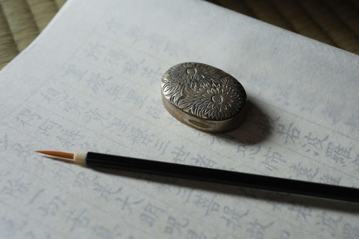 a silver bead with a floral design, a black pencil, and a piece of paper with Japanese characters on it.