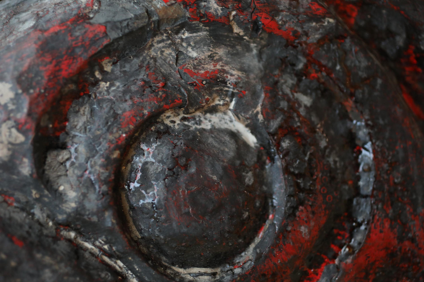 a close-up view of a textured surface with a mix of black, red, and gray colors. The surface appears to be a metal or concrete material, with a circular or oval shape in the center. The image does not contain any discernible text or specific objects, and the overall composition is simple and minimalistic.