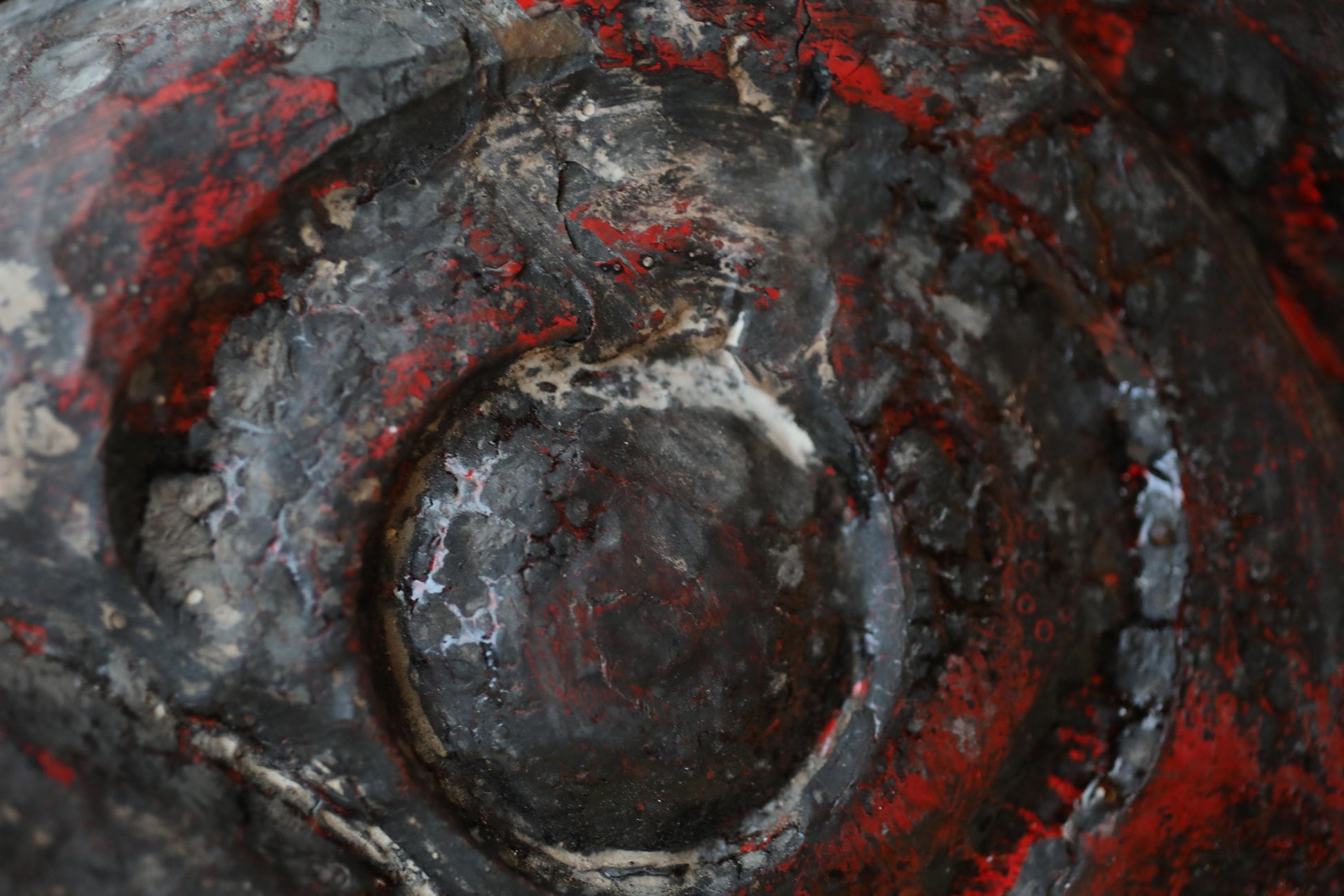 a close-up view of a textured surface with a mix of black, red, and gray colors. The surface appears to be a metal or concrete material, with a circular or oval shape in the center. The image does not contain any discernible text or specific objects, and the overall composition is simple and minimalistic.