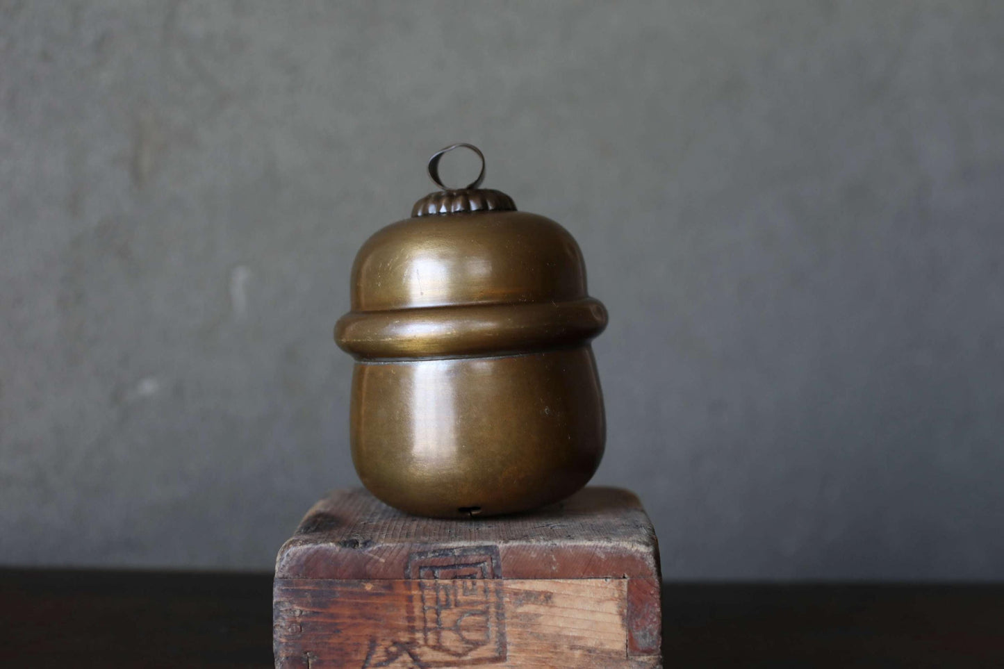 A brass container with a lid sits on a wooden crate against a gray background.
