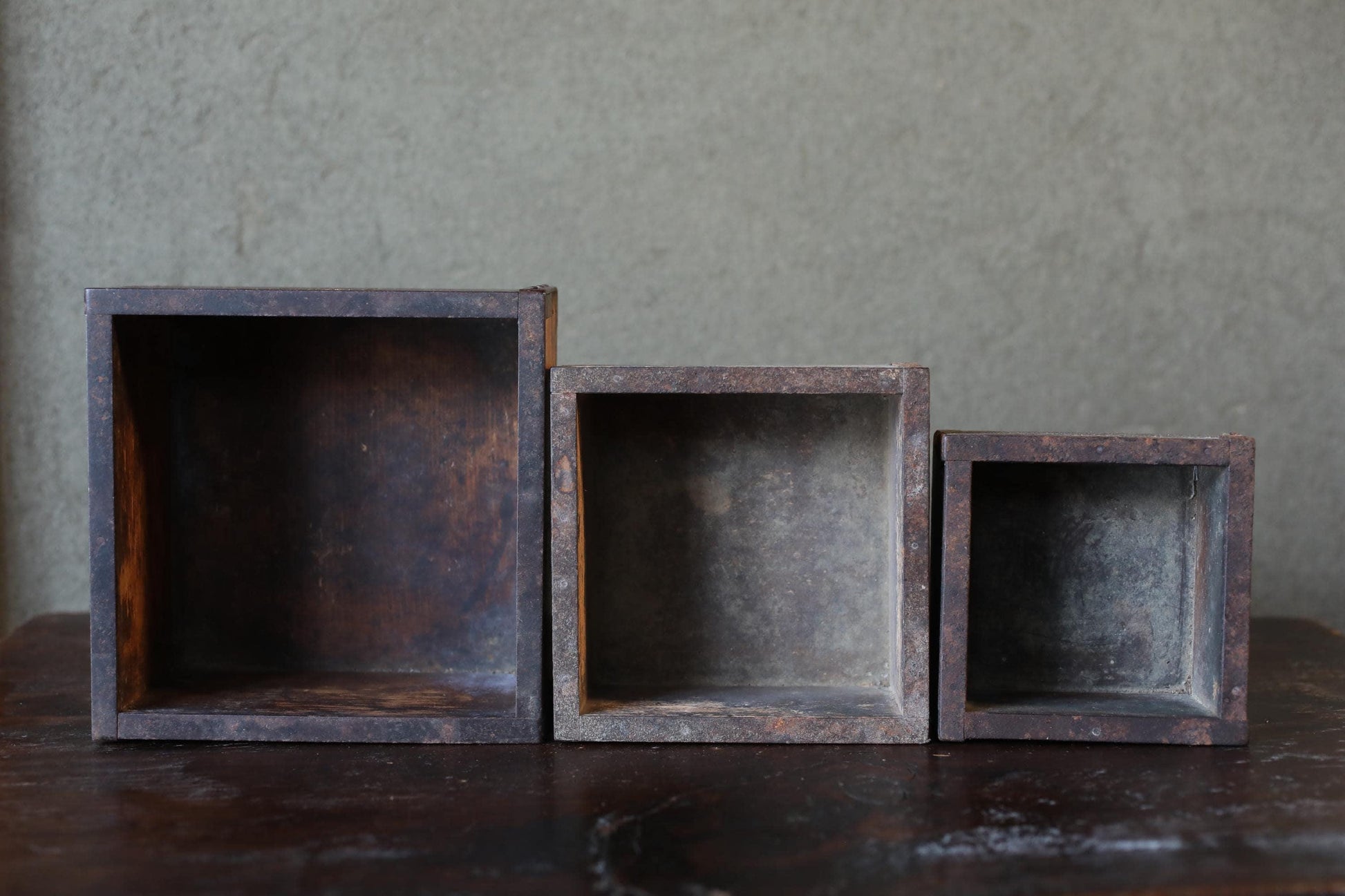 Three old, rusted metal boxes of varying sizes are arranged on a wooden surface against a plain gray wall.