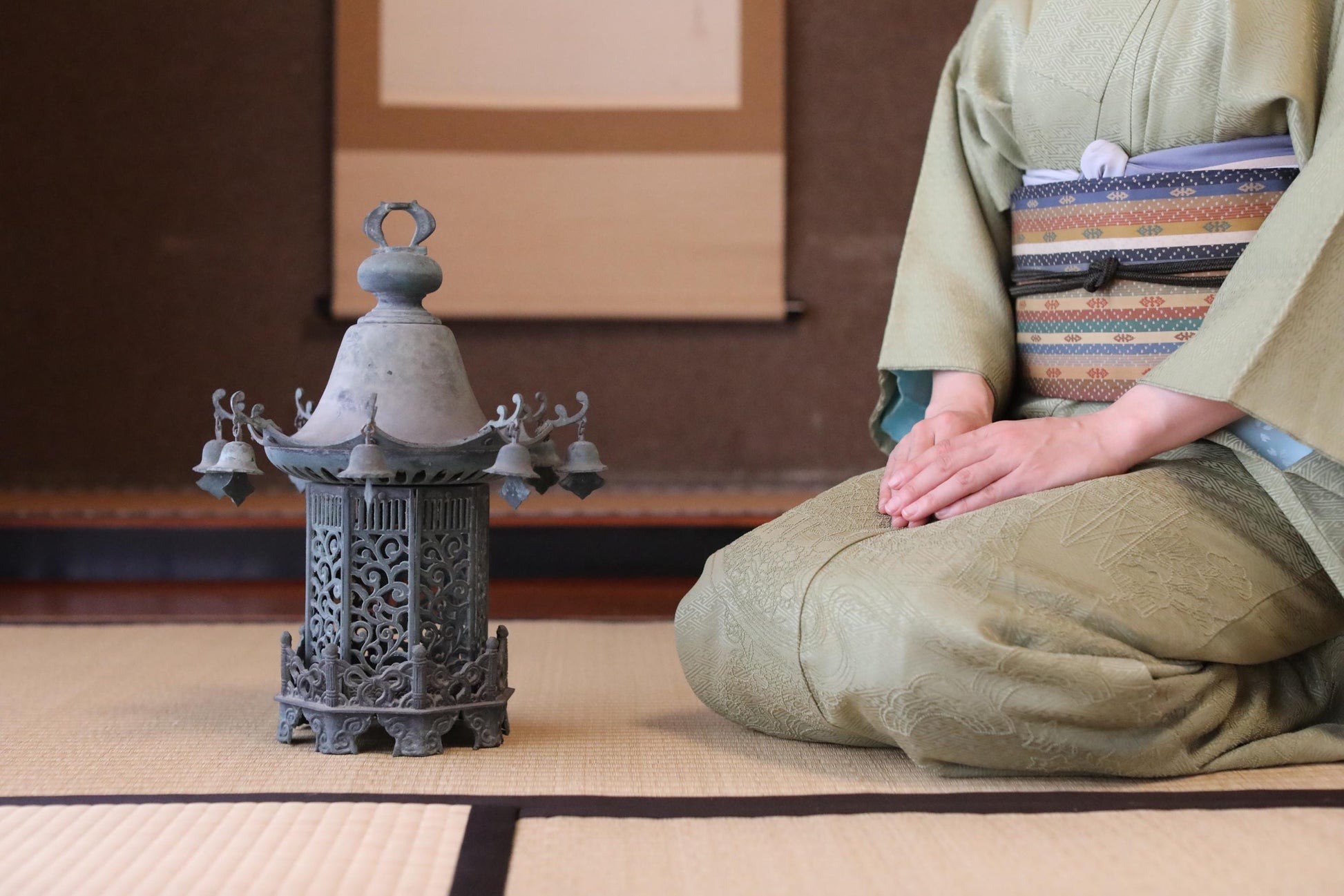 A person in traditional Japanese attire, including a kimono, is seated on the floor next to a decorative metal lantern.