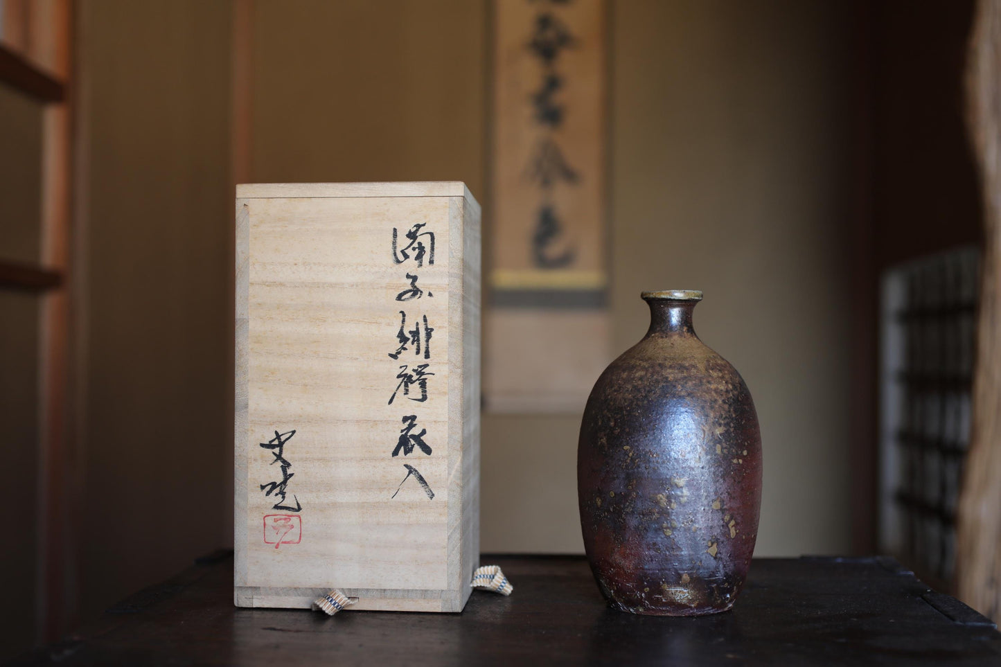 以下が含まれることがあります： 細い首の、素朴な濃い茶色の陶器の酒瓶と、日本の書道が書かれた木箱。ボトルは質感のある表面と金属製の縁があります。箱は明るい色で、黒インクで書かれています。