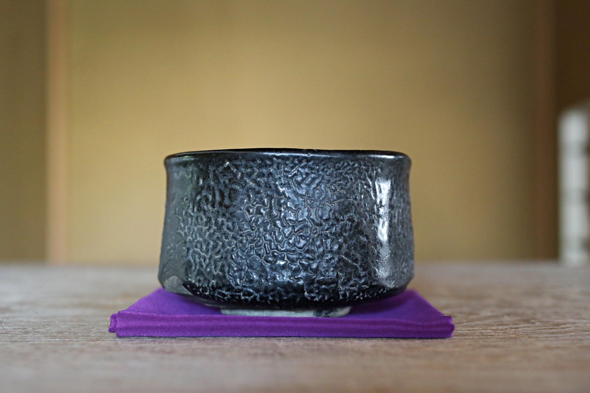 A black ceramic bowl with a textured surface sits on a purple coaster on a wooden table.