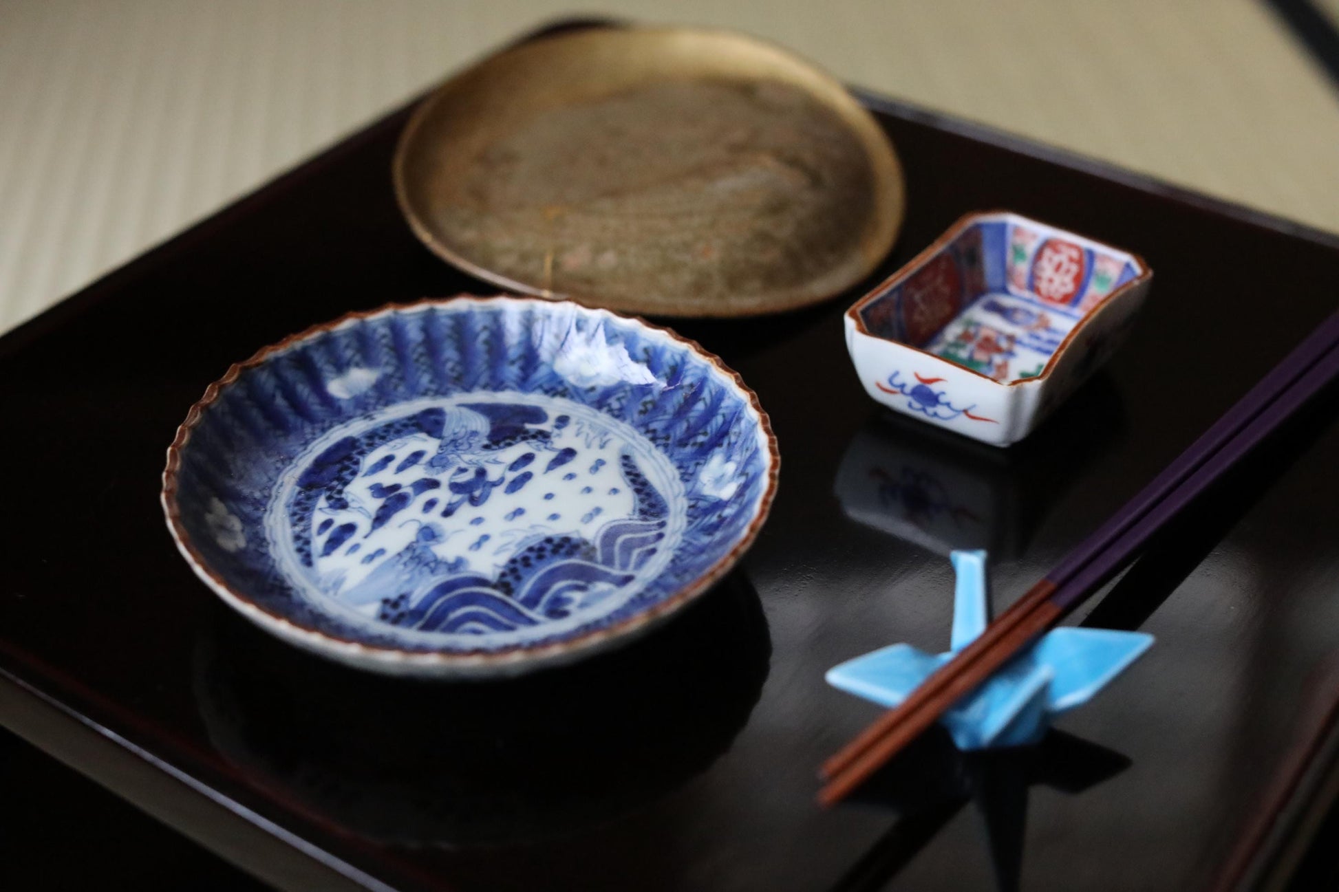 a variety of Japanese-style dishes and utensils, including a blue and white plate with an intricate design, a small bowl, and a pair of chopsticks, all arranged on a dark surface.