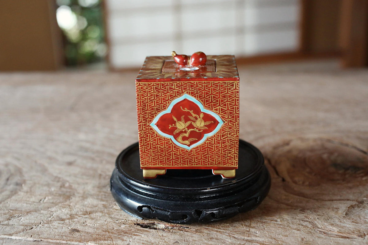 A small, ornate box with a red and gold design sits on a black stand on a wooden surface.