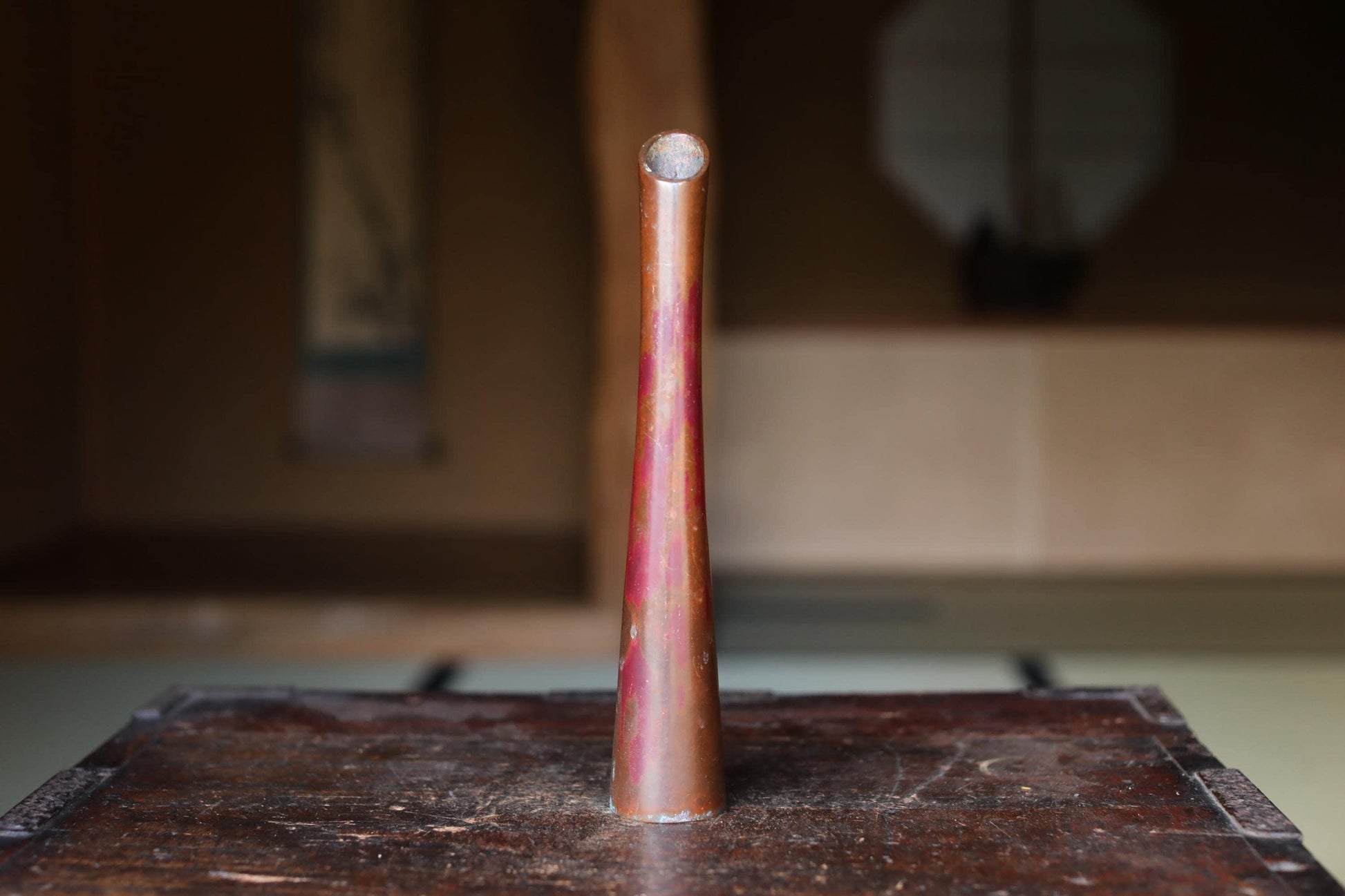 A tall, narrow copper vase with a pointed top sits on a wooden surface, with a blurred background suggesting an indoor setting.