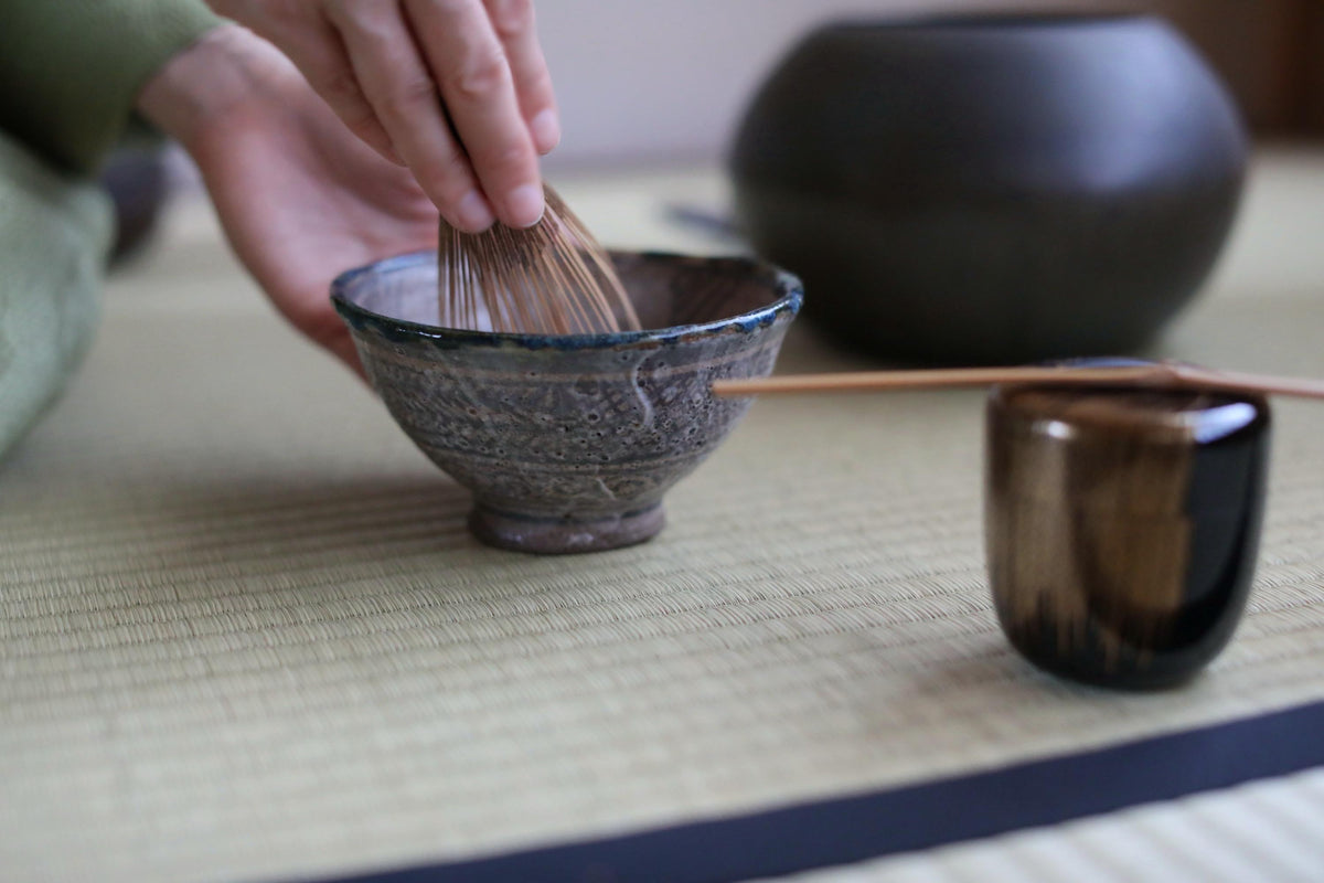 A person's hand is shown stirring a small bowl with a whisk, while a larger bowl and a small cup are visible in the background.
