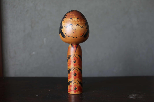 A wooden doll with a face and a floral pattern on its body, standing on a dark surface against a gray background.