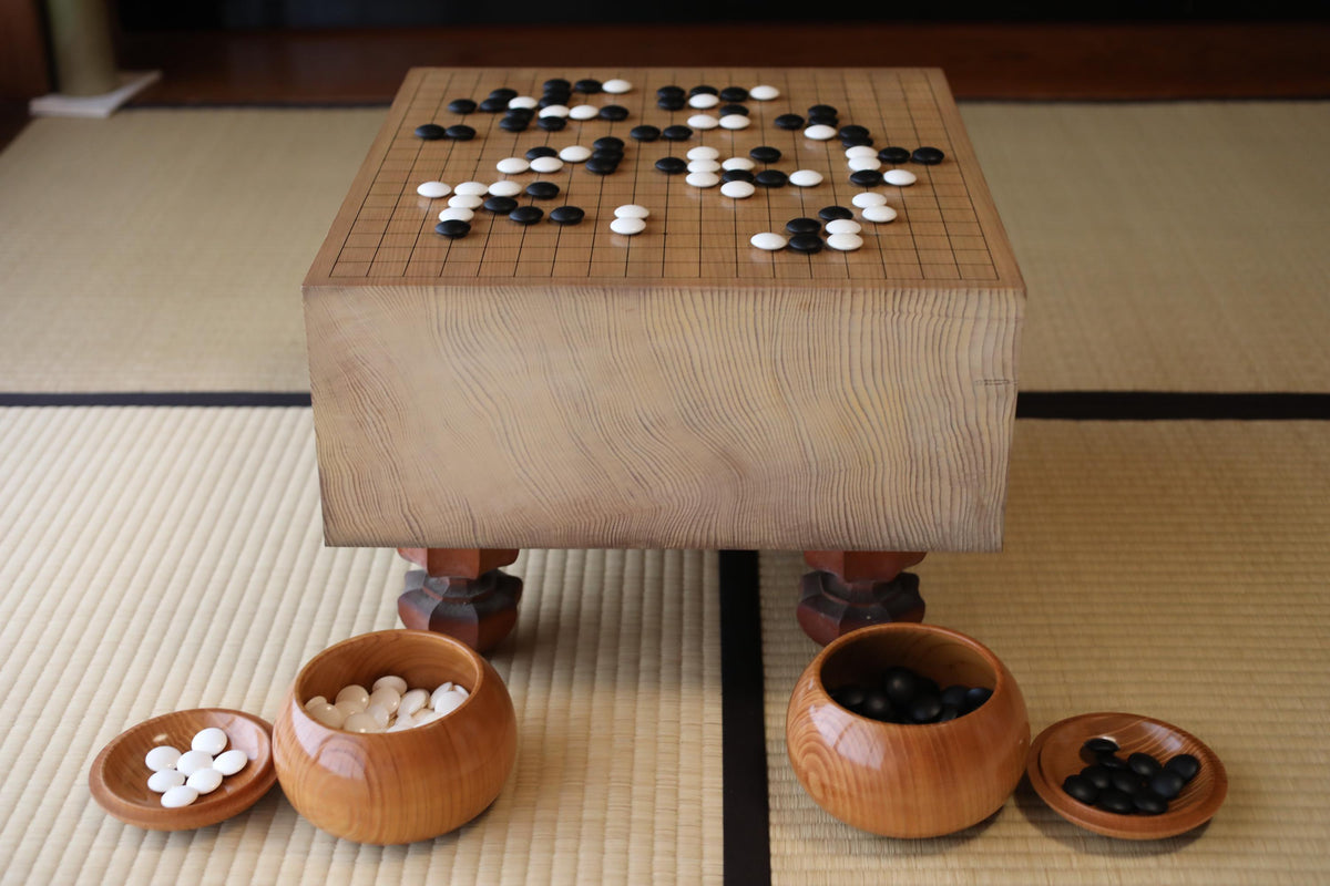 A wooden game board with a checkerboard pattern and a variety of small wooden bowls containing black and white objects, likely game pieces, are arranged on a tatami mat.