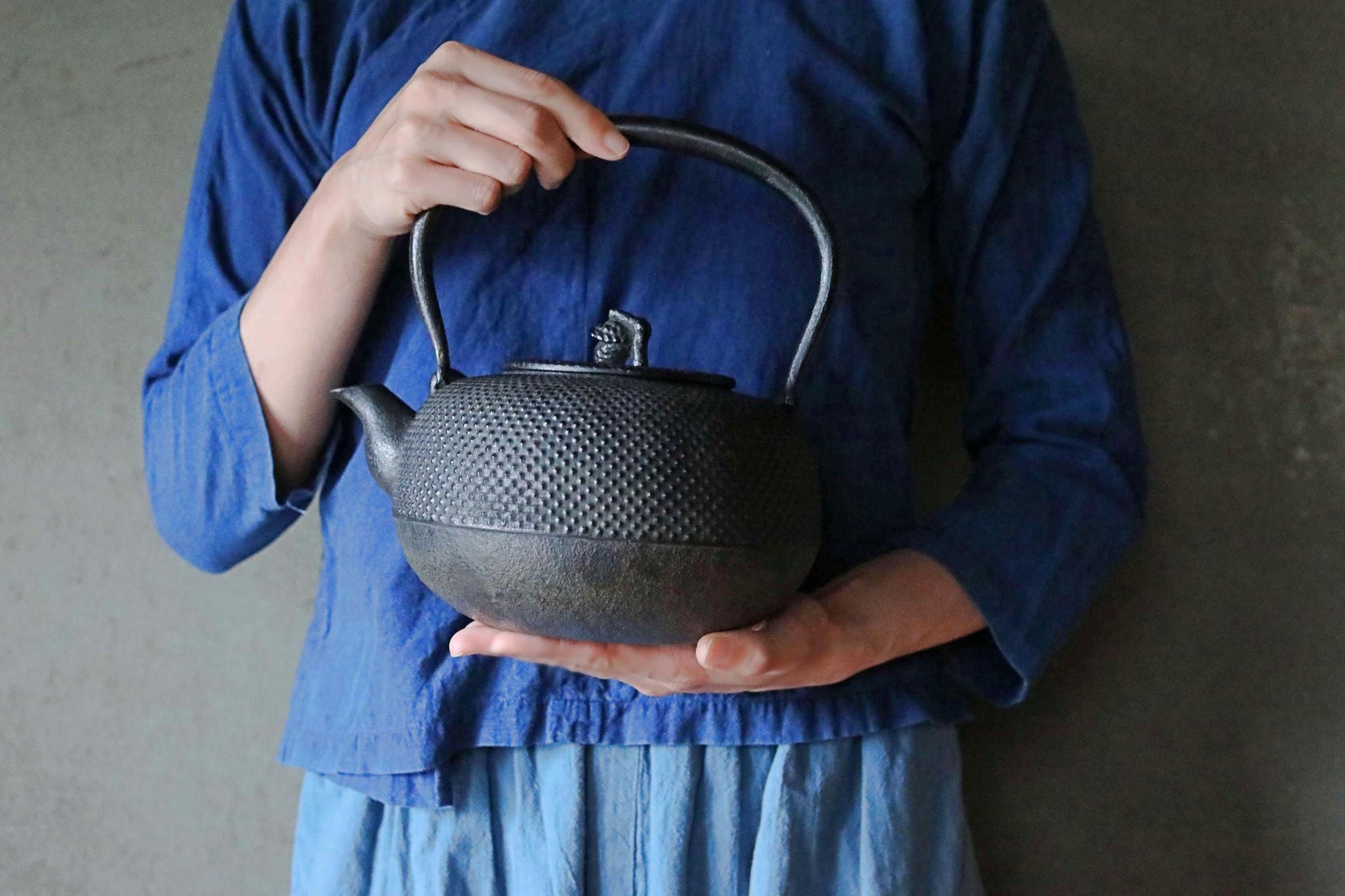 A person wearing a blue shirt and holding a black metal teapot.