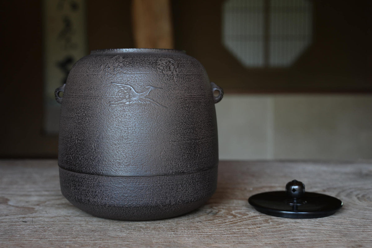 A black ceramic jar with a bird design sits on a wooden surface, with a black lid resting next to it.