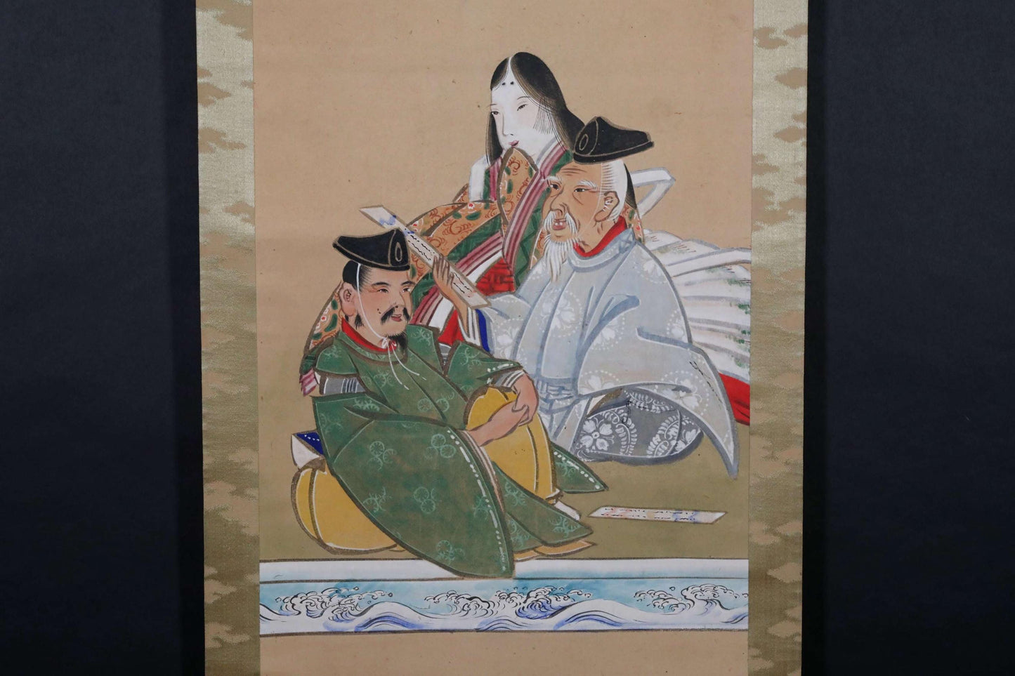 以下が含まれることがあります： 伝統的な衣装を着た3人の人物を描いた、ヴィンテージの日本の巻物絵画。人物は座っており、会話に夢中になっているように見える。絵画は、茶色、緑、青の落ち着いた色合いで描かれており、金色のアクセントが施されている。巻物はクリーム色の背景に飾られている。