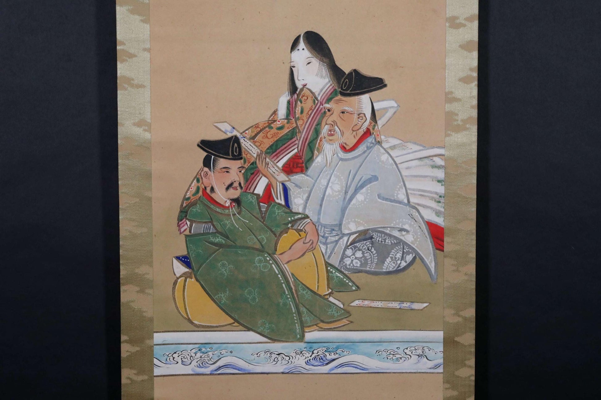 以下が含まれることがあります： 伝統的な衣装を着た3人の人物を描いた、ヴィンテージの日本の巻物絵画。人物は座っており、会話に夢中になっているように見える。絵画は、茶色、緑、青の落ち着いた色合いで描かれており、金色のアクセントが施されている。巻物はクリーム色の背景に飾られている。