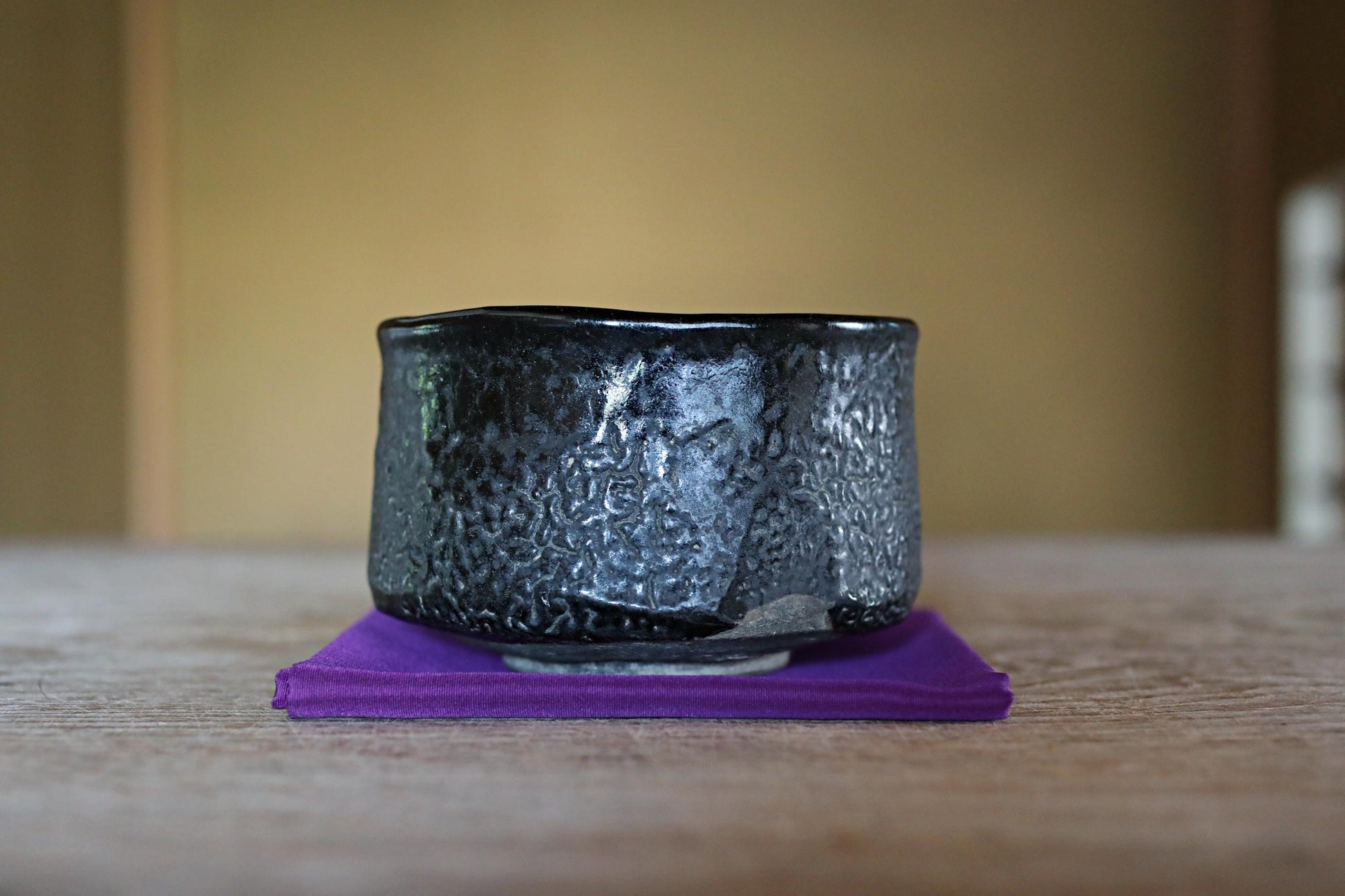 A black ceramic bowl with a textured surface sits on a purple coaster on a wooden table.