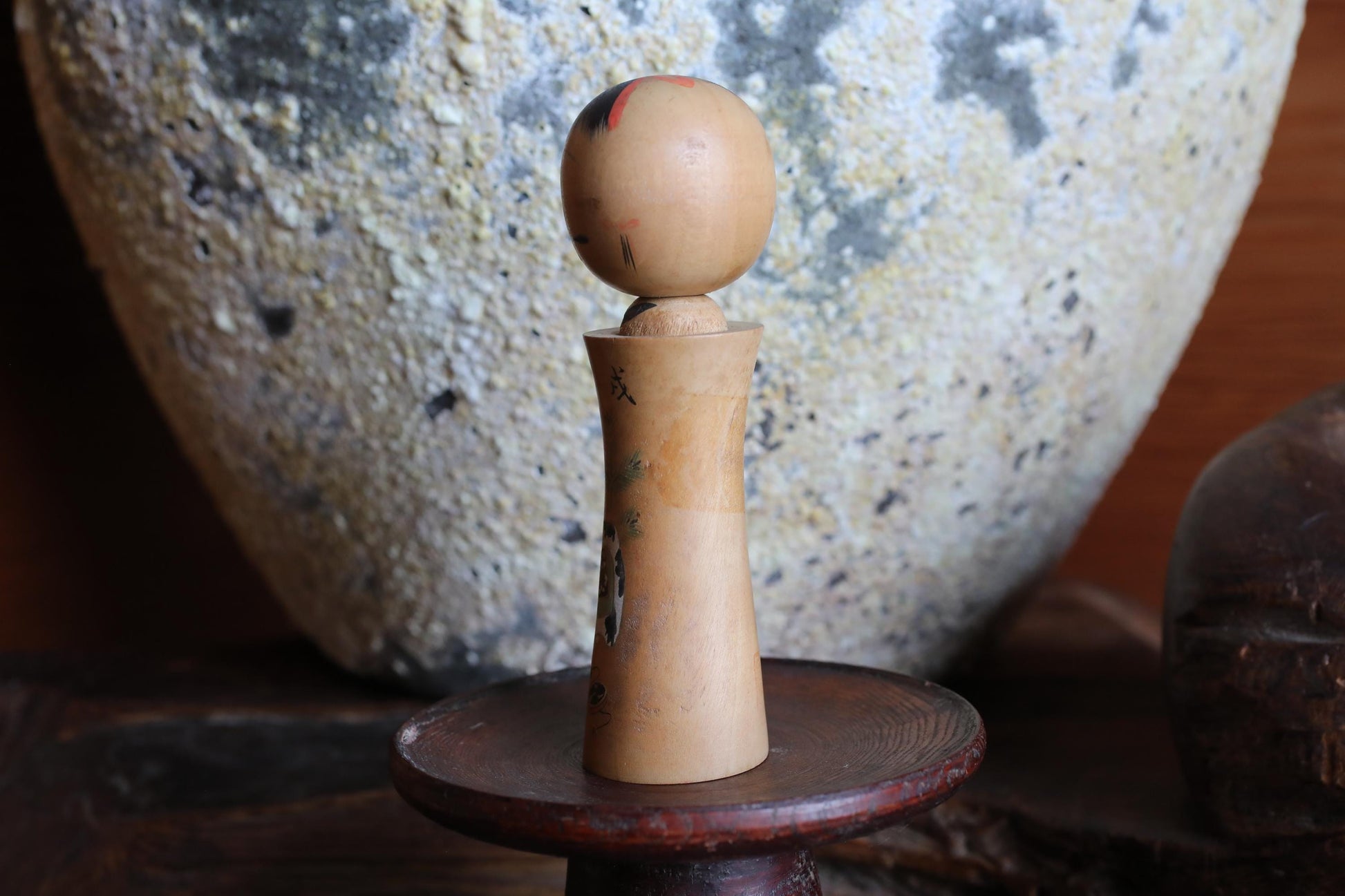 A wooden figurine with a round head and a small body is placed on a wooden stand in front of a large, textured stone vase.