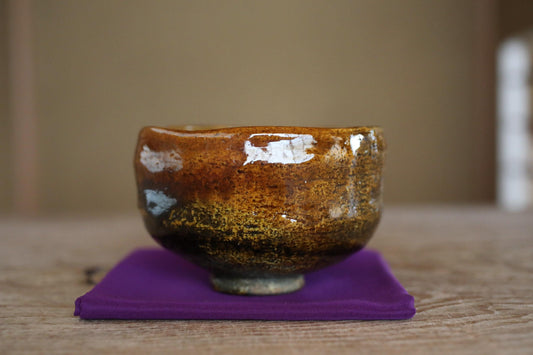 A brown ceramic bowl with a white speckled design sits on a purple cloth on a wooden surface.