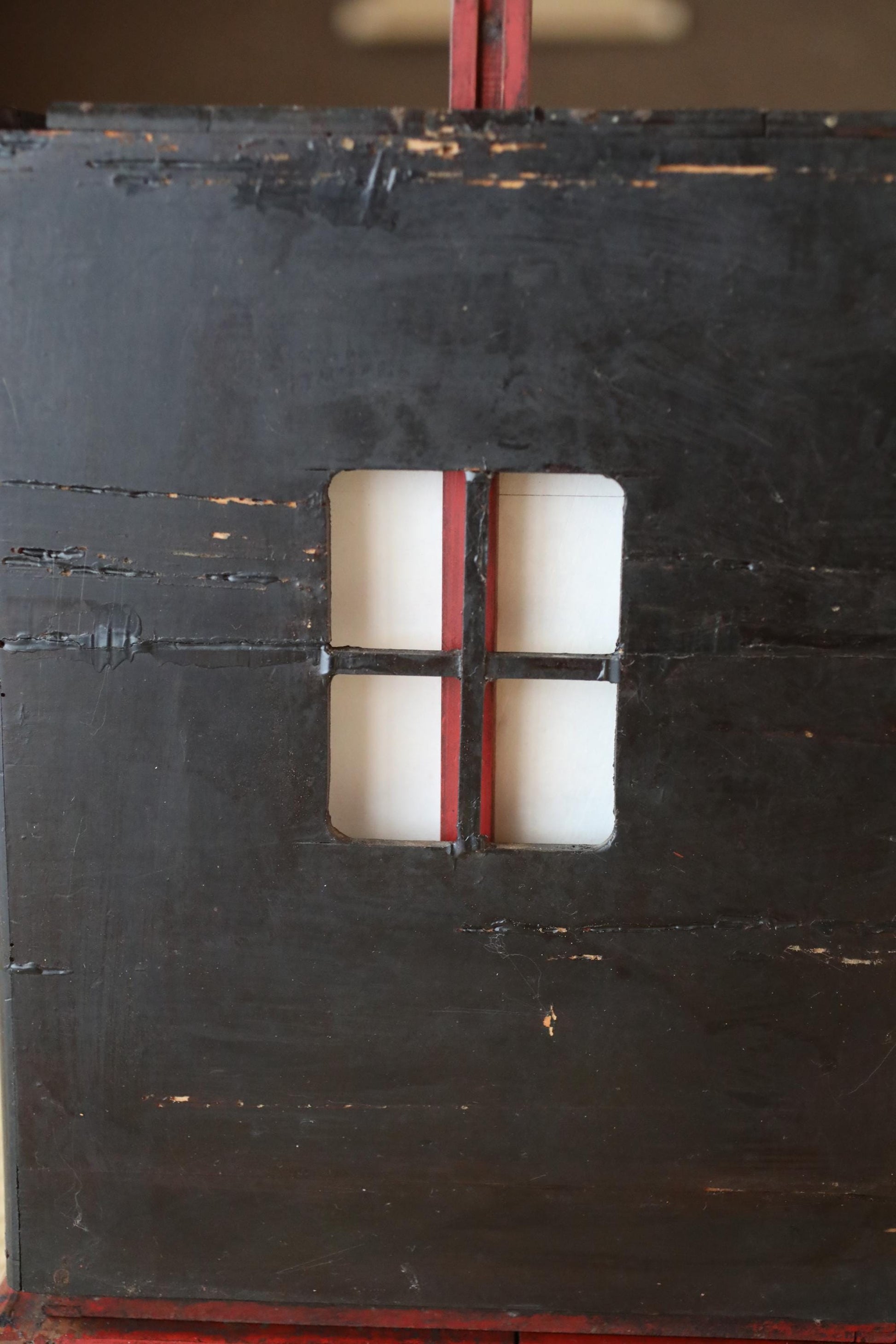 a close-up view of a black, weathered surface with a red and white striped pattern. There is a small, white window cut into the surface, which appears to be a part of a larger structure.
