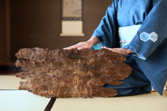 A person in a blue kimono is seated on a tatami mat, holding a large, intricately carved piece of driftwood.