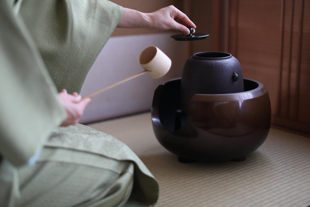 A person in a green robe is pouring tea from a black teapot into a brown teacup, with a wooden spoon in their hand.