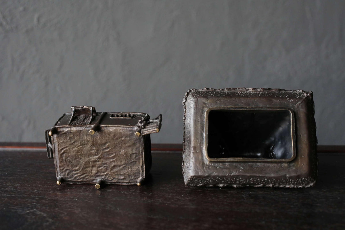 Two antique metal boxes, one square and one rectangular, are placed on a dark surface against a gray background.
