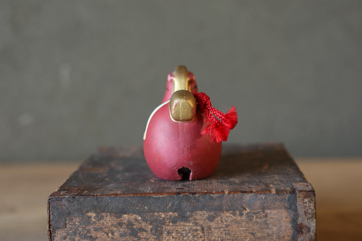 A red ceramic bird figurine with a gold handle and red string is displayed on a wooden surface against a gray background.