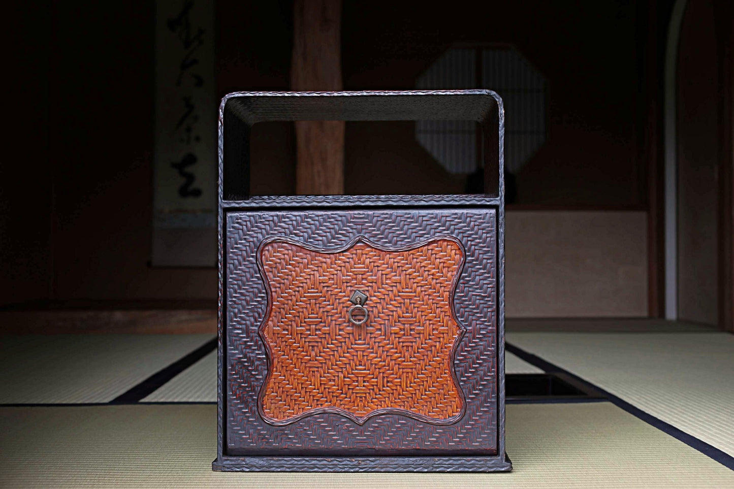 A traditional Japanese wooden cabinet with a woven design and a metal handle on the front, placed on a tatami mat in a dimly lit room.
