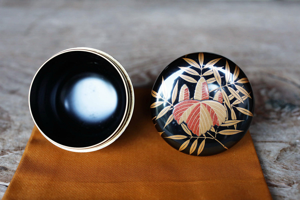 a black and gold decorative bowl with a floral design, placed on a wooden surface next to an open lid of the same bowl.