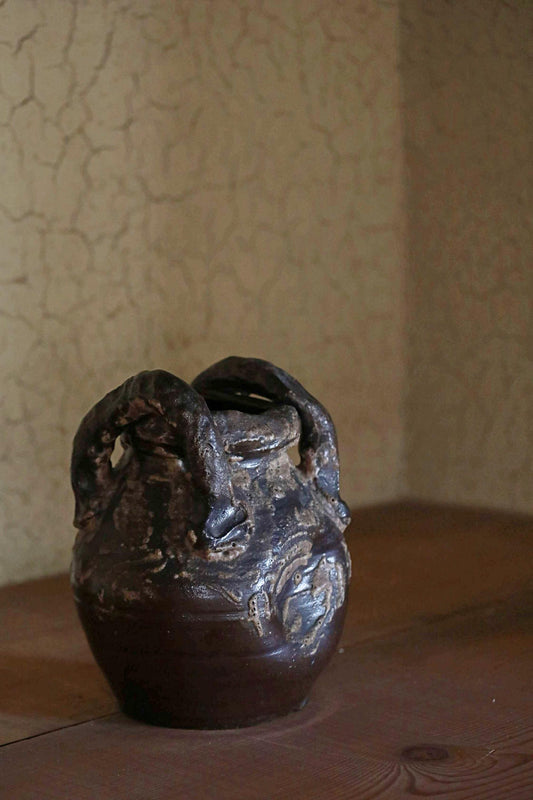 A dark-colored ceramic vase with a unique, sculptural design sits on a wooden surface against a textured wall.