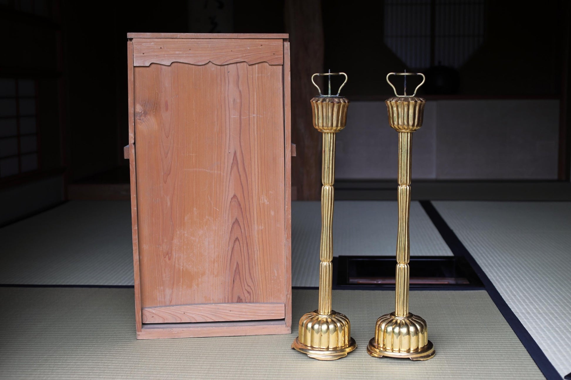a pair of golden candlesticks placed next to a wooden panel, both sitting on a tatami mat.