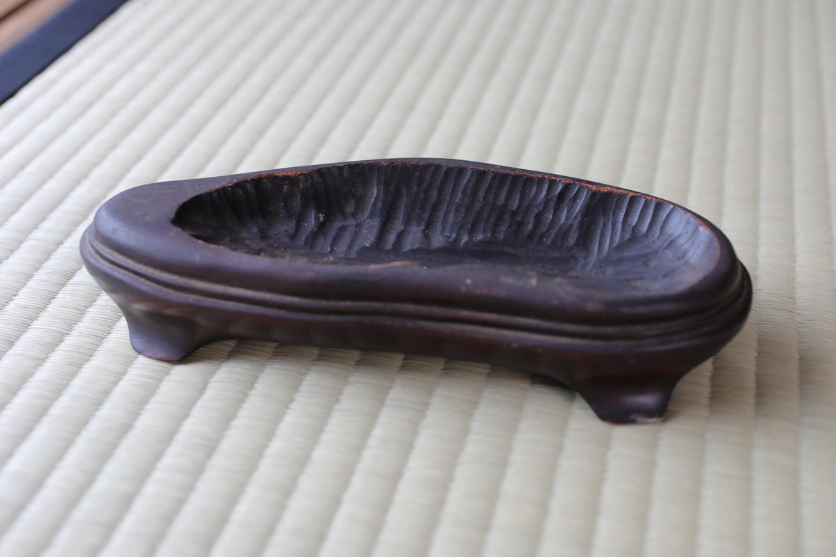 A dark brown, oval-shaped ceramic object with a textured surface, placed on a white surface with a woven texture.