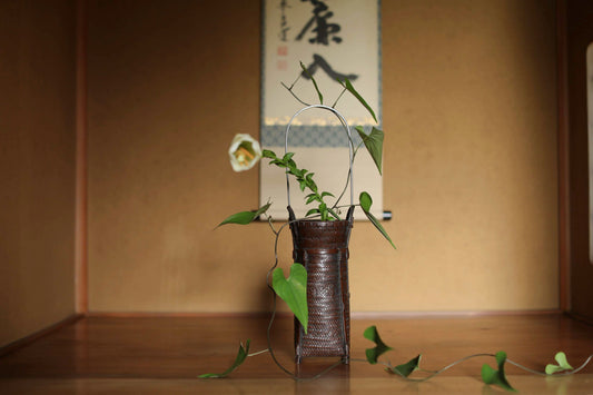 A small brown woven vase with a plant in it, placed on a wooden surface, with a traditional Japanese scroll hanging on the wall in the background.