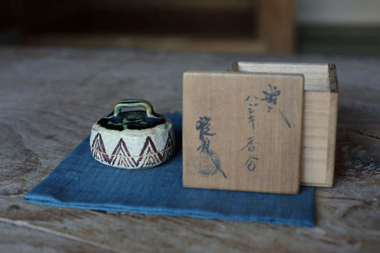 a ceramic object with a handle and a wooden box with Japanese characters on it, both placed on a blue cloth on a wooden surface.