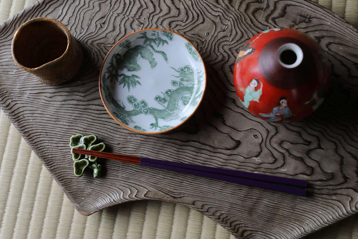 a variety of Japanese-style items arranged on a wooden surface, including a red vase, a green and white plate, a small ceramic cup, and a pair of chopsticks.