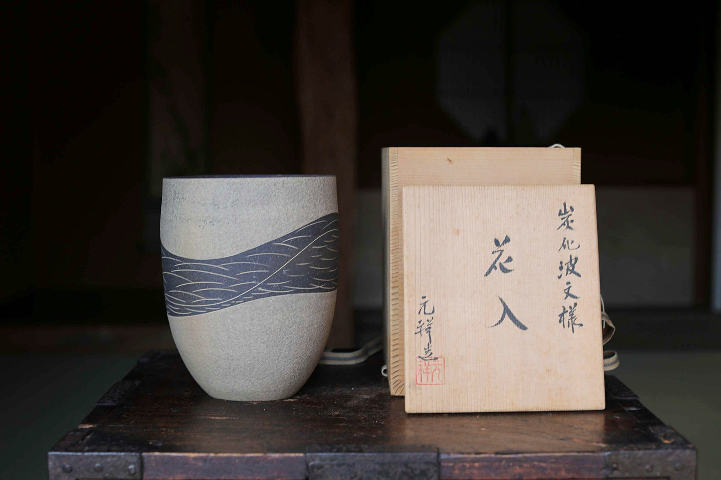 a ceramic vase with a wave pattern design, placed on a wooden surface next to a wooden box with Japanese characters on it.