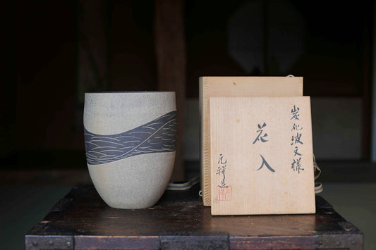 a ceramic vase with a wave pattern design, placed on a wooden surface next to a wooden box with Japanese characters on it.