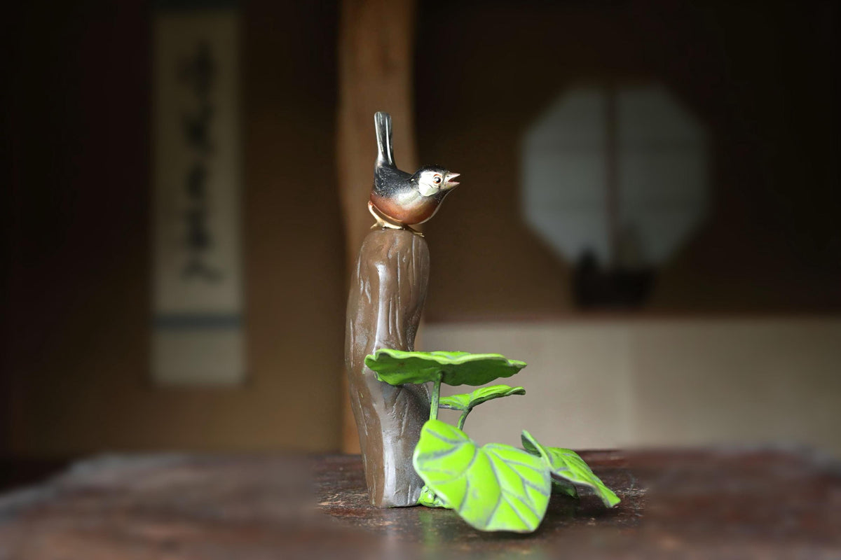 A small bird figurine perched on a branch with green leaves, set against a blurred background.