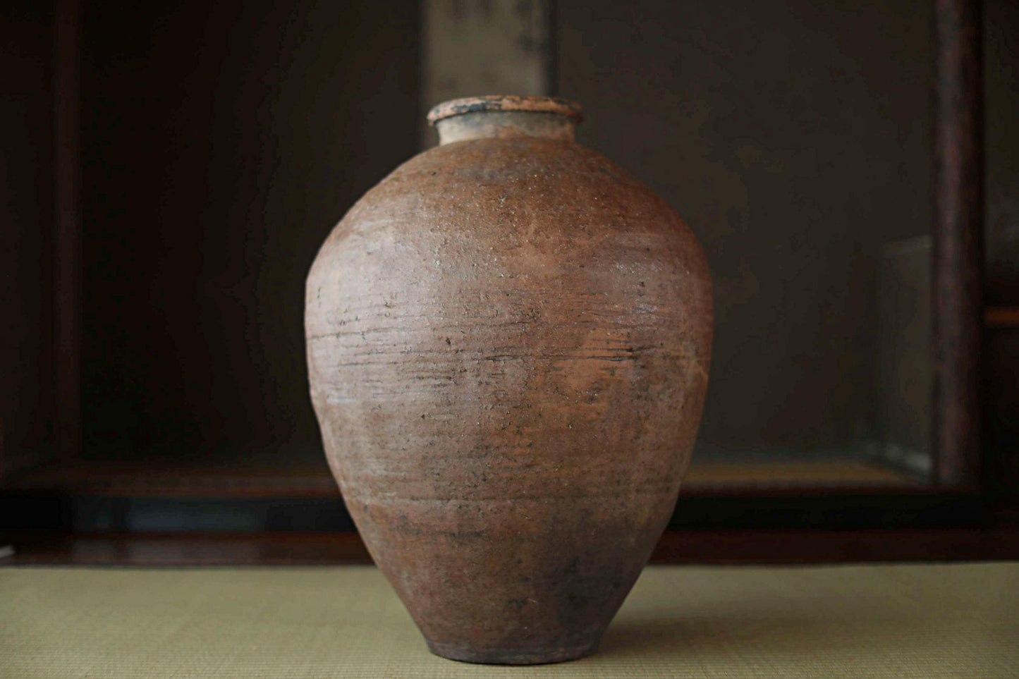 An old, weathered clay vase with a distinctive shape and texture, sitting on a surface against a dark background.