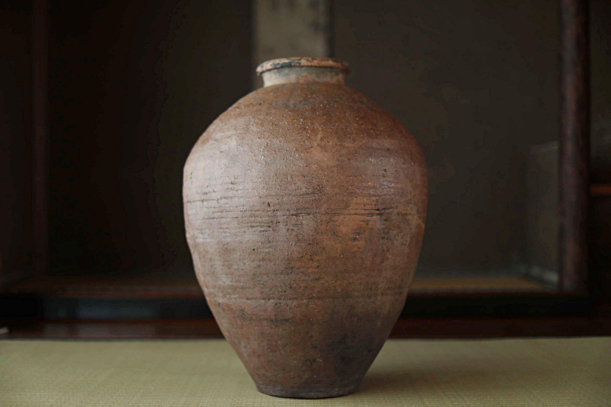 An old, weathered clay vase with a distinctive shape and texture, sitting on a surface against a dark background.