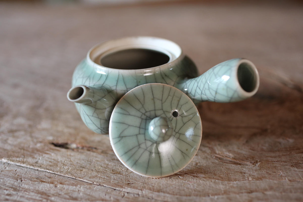 A ceramic teapot with a green and white color scheme, featuring a handle and spout, is placed on a wooden surface.