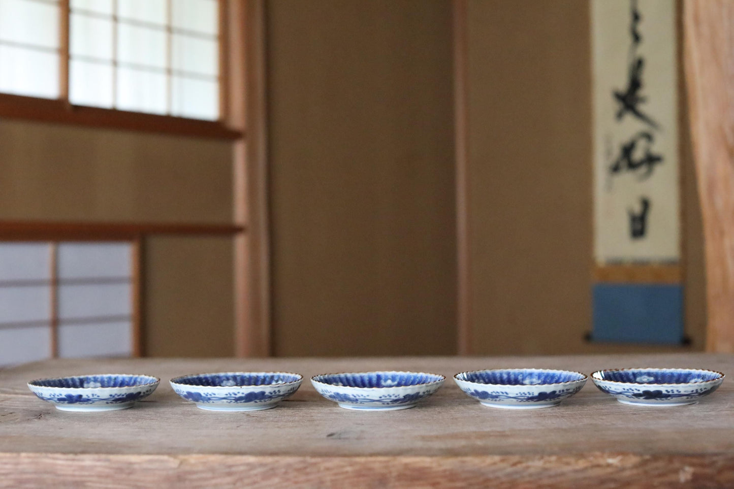 A row of blue and white ceramic bowls arranged on a wooden surface, with a blurred background that includes a window and a poster.