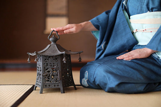 A person in traditional Japanese attire is seated on the floor, gently touching a small, ornate lantern with a black metal frame.