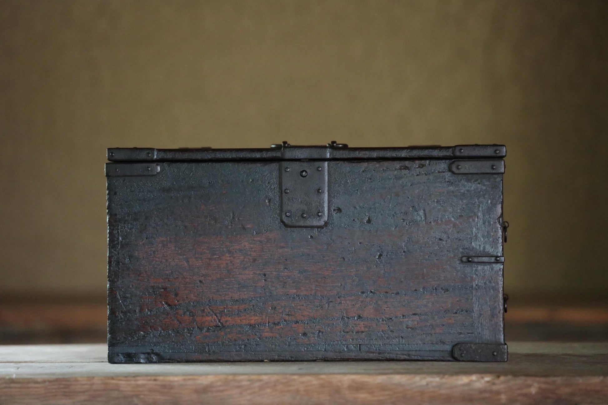 A dark wooden chest with metal clasps sits on a wooden surface against a plain background.