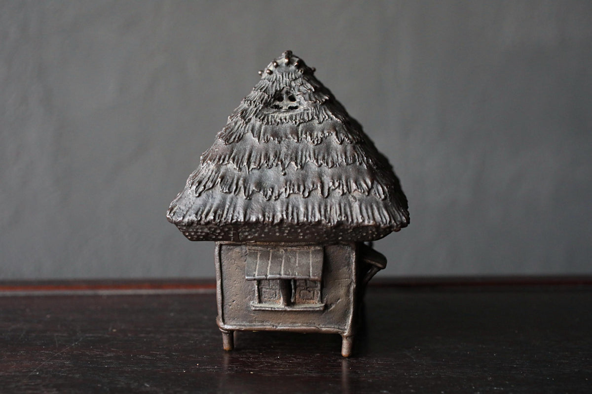 A small, intricately designed metal sculpture of a house with a pointed roof and a small window on the side, sitting on a dark surface against a gray background.